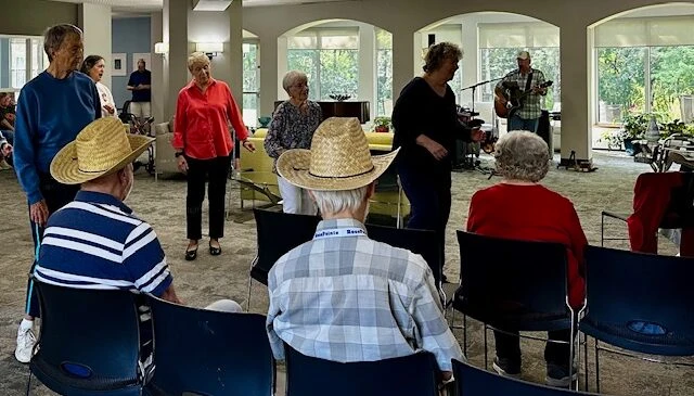 Seniors gathered in a community room enjoying live music, some seated and some standing with straw hats.