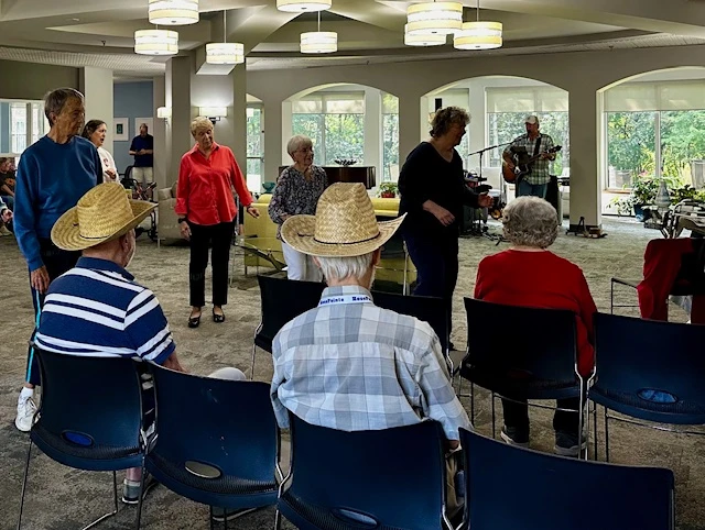 Seniors gathered in a community room enjoying live music, some seated and some standing with straw hats.